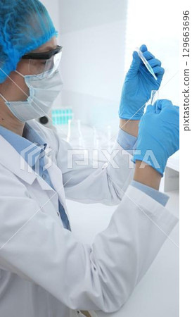 Medical science researcher wearing blue protective gear of cap, gloves and googles, is examining liquid in test tube in laboratory workspace. Medicine and health care concept 129663696