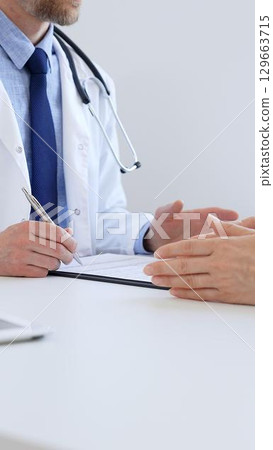 Doctor man taking notes while listening to a female patient explaining her symptoms during a medical consultation in the clinic office. Medicine and health care concept Doctor man taking notes while listening to a female patient explaining her symptoms during a medical consultation in the clinic office. Medicine and health care concept 129663715