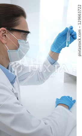Female researcher examining clear liquid in test tube, sitting near microscopes and lab equipment within sterile scientific workspace. Medicine, healthcare and science concept 129663722