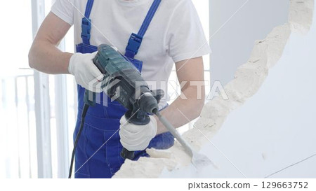 Male construction worker demolishing white wall with rotary hammer drill, wearing blue overalls and protective white gloves, closeup view 129663752