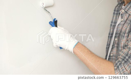 Male painter's gloved hand gripping paint roller, spreading white paint across wall during home interior transformation with professional precision, close-up view. Renovation concept 129663755