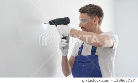 Middle aged man wearing blue overall, is drilling hole in white wall, wearing protective gear during home improvement project with cordless power tool Middle aged man wearing blue overall, is drilling hole in white wall, wearing protective gear during home improvement project with cordless power tool 129663757