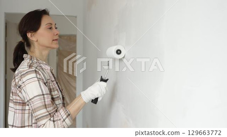 Painter woman wearing gloves is skillfully applying paint to a white wall with a roller in an apartment undergoing renovation, transforming the space with her creative touch 129663772