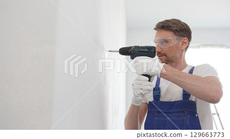 Middle aged male construction worker in a blue jumpsuit, safety goggles, and work gloves is screwing a screw into the wall using a cordless screwdriver, carrying out a careful home renovation with 129663773