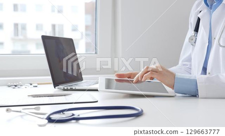 Female doctor wearing lab coat and stethoscope using digital tablet with laptop and medical records on desk in medical office, showcasing modern healthcare technology. Medicine concept 129663777