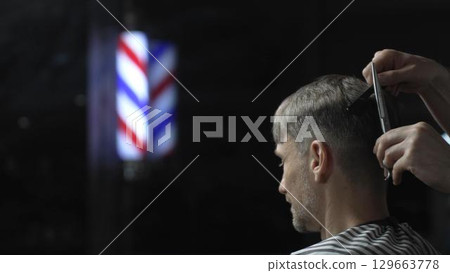 Professional barber cutting hair of male client in barbershop, using scissors and comb, with barber pole in background, creating stylish and modern haircut Professional barber cutting hair of male client in barbershop, using scissors and comb, with barber pole in background, creating stylish and modern haircut 129663778
