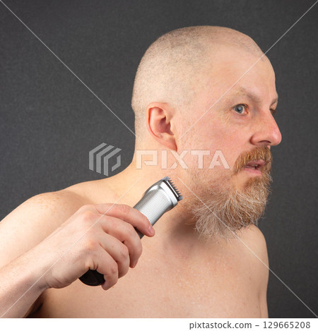 Man grooming his beard with an electric trimmer in a minimalistic setting 129665208