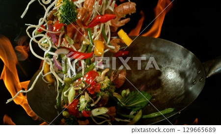 Closeup of chef throwing asian vegetable with meat mix from wok pan in fire. Fresh asian food preparation on black background. Closeup of chef throwing asian vegetable with meat mix from wok pan in fire. Fresh asian food preparation on black background. 129665689