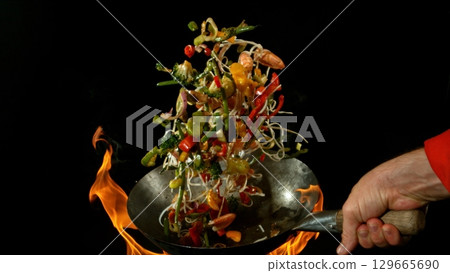 Closeup of chef throwing asian vegetable with meat mix from wok pan in fire. Fresh asian food preparation on black background. 129665690