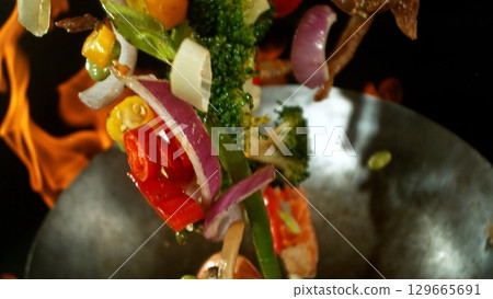 Closeup of chef throwing asian vegetable with meat mix from wok pan in fire. Fresh asian food preparation on black background. Closeup of chef throwing asian vegetable with meat mix from wok pan in fire. Fresh asian food preparation on black background. 129665691