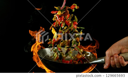 Closeup of chef throwing asian vegetable with meat mix from wok pan in fire. Fresh asian food preparation on black background. 129665693