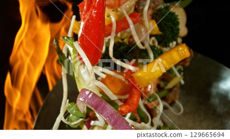 Closeup of chef throwing asian vegetable with meat mix from wok pan in fire. Fresh asian food preparation on black background. Closeup of chef throwing asian vegetable with meat mix from wok pan in fire. Fresh asian food preparation on black background. 129665694
