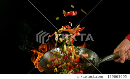 Closeup of chef throwing asian vegetable with meat mix from wok pan in fire. Fresh asian food preparation on black background. Closeup of chef throwing asian vegetable with meat mix from wok pan in fire. Fresh asian food preparation on black background. 129665695