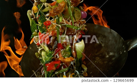 Closeup of chef throwing asian vegetable with meat mix from wok pan in fire. Fresh asian food preparation on black background. Closeup of chef throwing asian vegetable with meat mix from wok pan in fire. Fresh asian food preparation on black background. 129665699