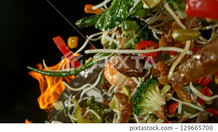 Closeup of chef throwing asian vegetable with meat mix from wok pan in fire. Fresh asian food preparation on black background. Closeup of chef throwing asian vegetable with meat mix from wok pan in fire. Fresh asian food preparation on black background. 129665700