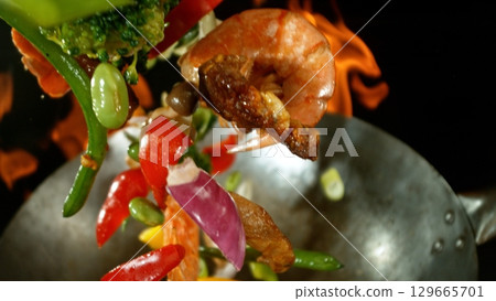 Closeup of chef throwing asian vegetable with meat mix from wok pan in fire. Fresh asian food preparation on black background. Closeup of chef throwing asian vegetable with meat mix from wok pan in fire. Fresh asian food preparation on black background. 129665701