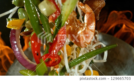 Closeup of chef throwing asian vegetable with meat mix from wok pan in fire. Fresh asian food preparation on black background. Closeup of chef throwing asian vegetable with meat mix from wok pan in fire. Fresh asian food preparation on black background. 129665703