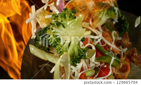 Closeup of chef throwing asian vegetable with meat mix from wok pan in fire. Fresh asian food preparation on black background. Closeup of chef throwing asian vegetable with meat mix from wok pan in fire. Fresh asian food preparation on black background. 129665704
