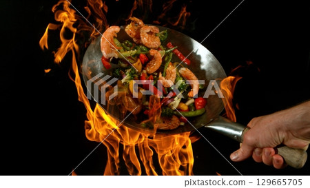 Closeup of chef throwing asian vegetable with meat mix from wok pan in fire. Fresh asian food preparation on black background. Closeup of chef throwing asian vegetable with meat mix from wok pan in fire. Fresh asian food preparation on black background. 129665705