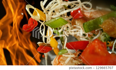 Closeup of chef throwing asian vegetable with meat mix from wok pan in fire. Fresh asian food preparation on black background. Closeup of chef throwing asian vegetable with meat mix from wok pan in fire. Fresh asian food preparation on black background. 129665707