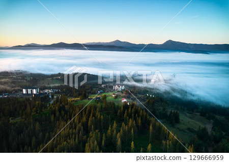 Aerial view of misty morning in mountains, with small village nestled among trees. Fog gently blankets landscape, creating serene and picturesque scene, as sun begins to rise in the background. 129666959