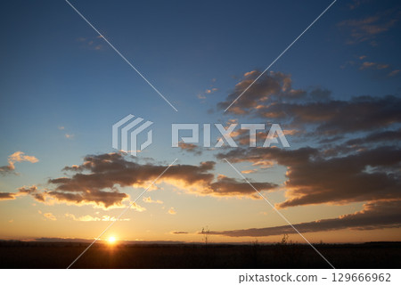 Sun sets low on horizon, casting golden glow across landscape. Fluffy clouds, illuminated with warm hues, float against clear blue sky. Serene scene tranquil, with last light of day fading gently. Sun sets low on horizon, casting golden glow across landscape. Fluffy clouds, illuminated with warm hues, float against clear blue sky. Serene scene tranquil, with last light of day fading gently. 129666962