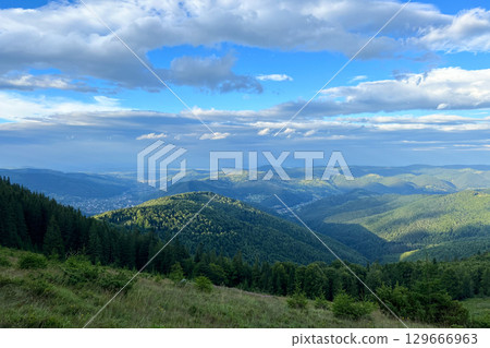 Breathtaking view of valley nestled among rolling hills in Ukraine. Lush forested landscape stretches out to distant town Yaremche, bathed in soft sunlight under serene, cloud-speckled sky. 129666963