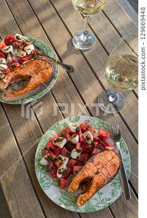Sunlit shot of two plates of grilled salmon steaks with caprese salad on wooden table. Two glasses of white wine are also visible, suggesting an outdoor summer meal Sunlit shot of two plates of grilled salmon steaks with caprese salad on wooden table. Two glasses of white wine are also visible, suggesting an outdoor summer meal 129669448