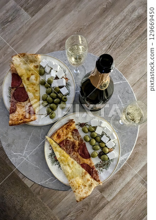 Top-down of two plates of pizza, each with two slices, served with green olives and feta cheese. Bottle of champagne and two glasses are on marble table, suggesting cozy, relaxed meal 129669450