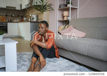 Young woman feeling down sitting on the floor in her living room 129669598