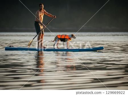 Man paddling on a stand-up paddleboard with dog wearing life jacket in calm water 129669922