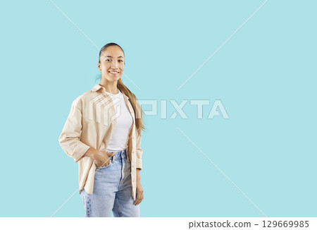 Young smiling female model posing in studio, standing with hand in pocket on azure blue background. 129669985
