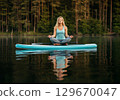 Woman practicing yoga on paddleboard in serene lake surrounded by lush trees and calm water 129670047