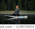 Woman practicing meditation on a paddleboard in tranquil lake surrounded by lush greenery 129670054