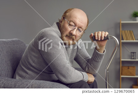 Sad, thoughtful, red-haired, elderly man with beard and glasses is sitting in gray armchair at home. 129670065
