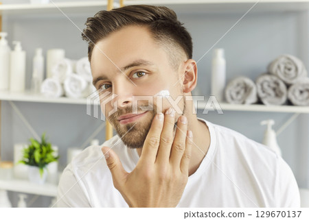 Handsome man smiling looking at camera, mirror, applying cream to cheek, hand gently touching face 129670137