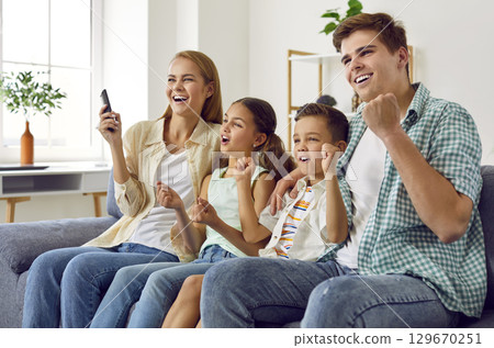 Cheerful family with children cheering together while watching TV at home in living room. 129670251