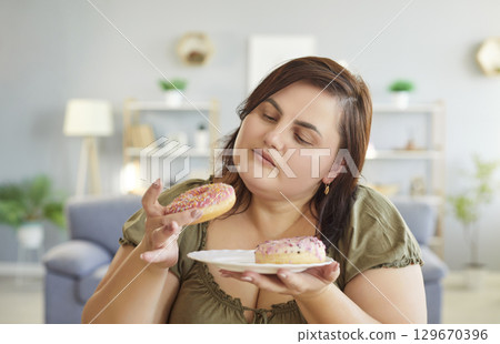 Overweight young fat woman sitting in living room and looking at sweet donut on plate. 129670396