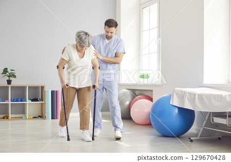 Physiotherapist Guiding Elderly Patient With Crutches During Rehabilitation 129670428