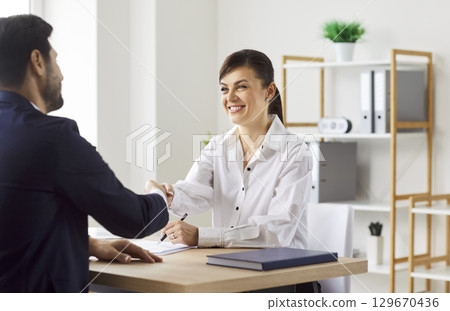Businesswoman shaking client hand after successful deal, sitting at desk in office, satisfied hr  129670436