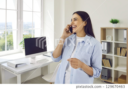 Happy smiling woman standing in office near workplace and talking on mobile phone. 129670463