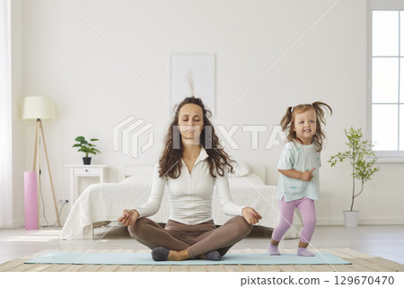 Relaxed mother practicing yoga in lotus pose with closed eyes while playful daughter running around. 129670470
