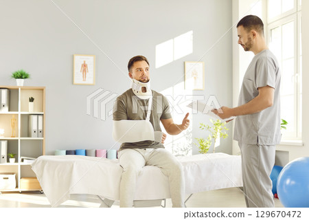 Doctor traumatologist examining man in neck brace with broken arm in medical office in exam room. Doctor traumatologist examining man in neck brace with broken arm in medical office in exam room. 129670472