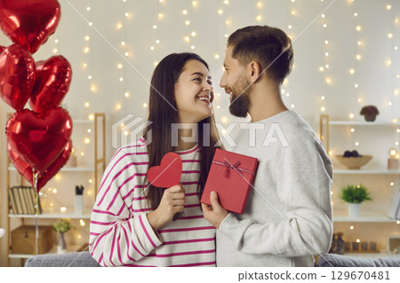 Happy smiling couple in love in romantic atmosphere in each other's arms, holding red heart and gift box Happy smiling couple in love in romantic atmosphere in each other's arms, holding red heart and gift box 129670481