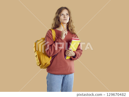 Half-turned thoughtful young girl student with backpack and books holding pencil in hands. 129670482