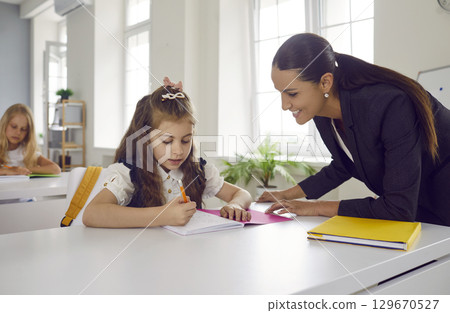 Smiling friendly teacher helping young schoolgirl in classroom Smiling friendly teacher helping young schoolgirl in classroom 129670527