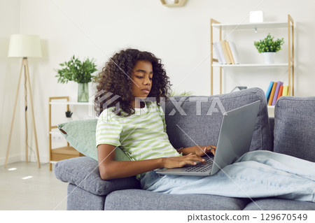 Serious young african teenager girl using laptop lying on sofa at home. Serious young african teenager girl using laptop lying on sofa at home. 129670549