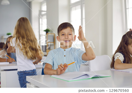 Portrait of small active schoolboy who raises his hand in class to ask question or answer. Portrait of small active schoolboy who raises his hand in class to ask question or answer. 129670560
