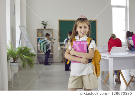 Portrait of a happy school girl with her notebooks and backpack standing in the classroom Portrait of a happy school girl with her notebooks and backpack standing in the classroom 129670580
