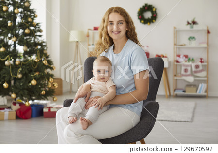 Portrait of happy mother with toddler sitting in festive room by Christmas tree during New Year eve. 129670592